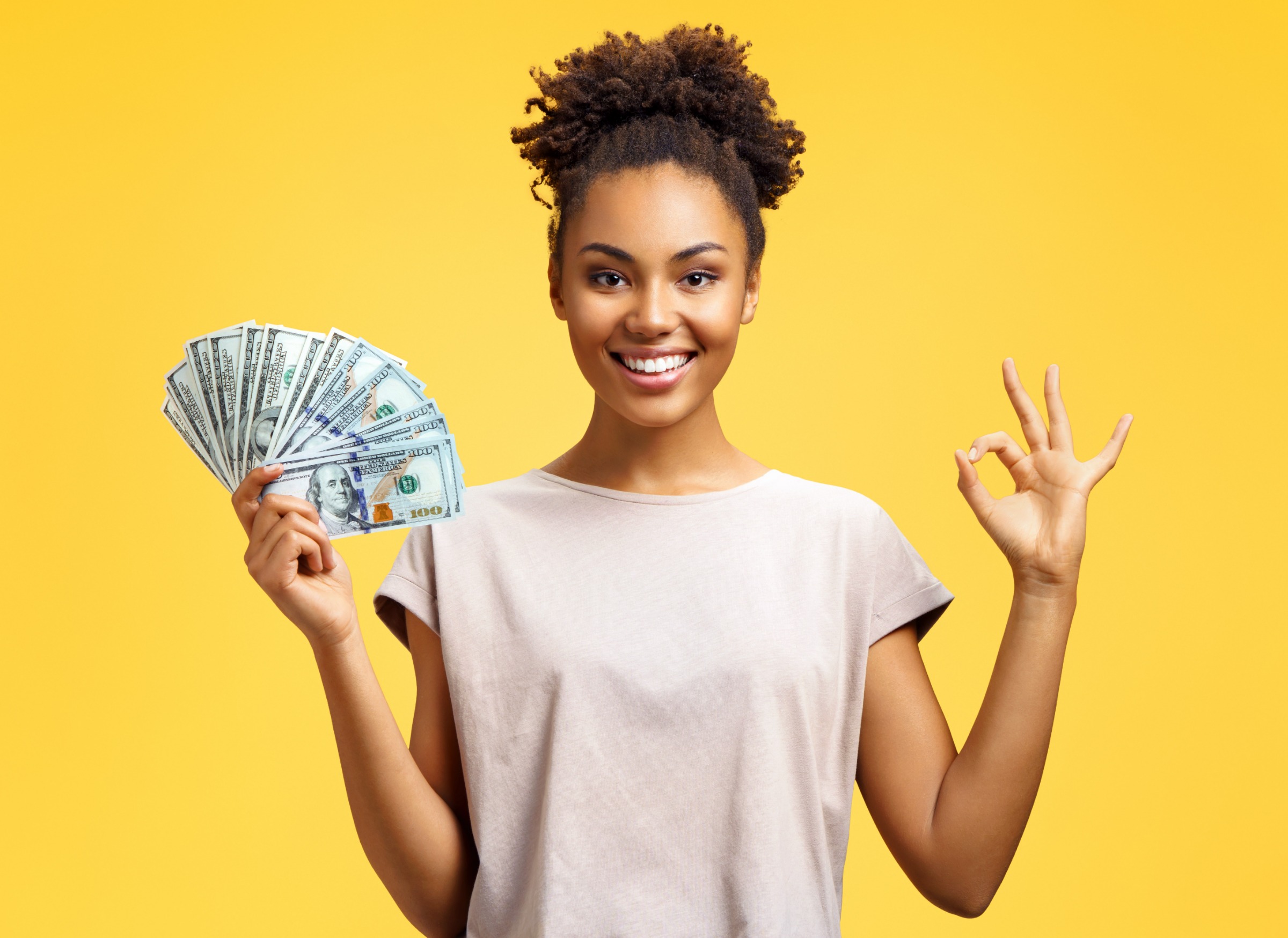 Smiling girl shows money cash and excellent sign, demonstrates that everything is fine. Photo of african american girl wears casual outfit on yellow background. Emotions and pleasant feelings concept.